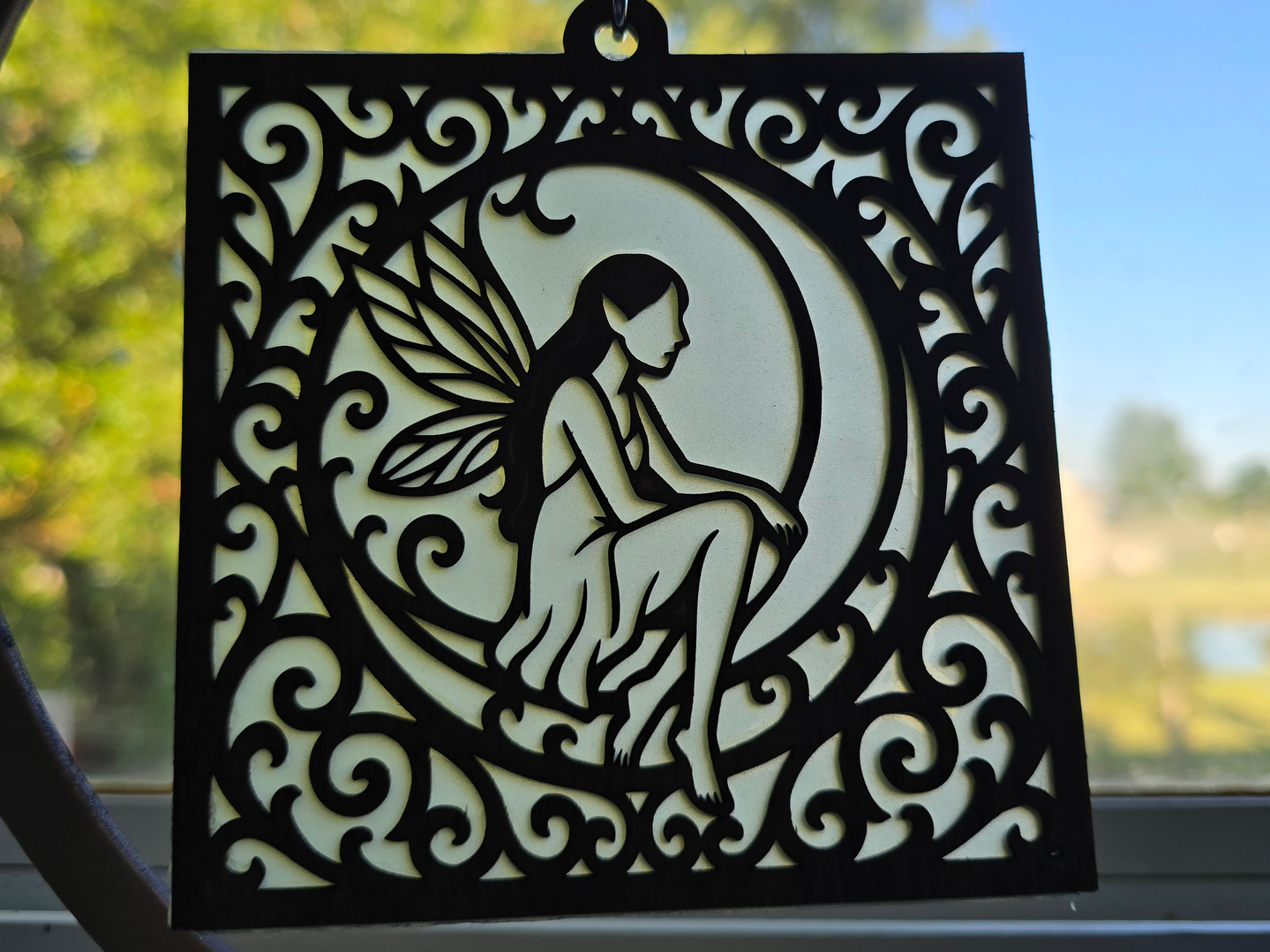 A laser-cut fairy sitting on a crescent moon inside an ornate sun catcher frame