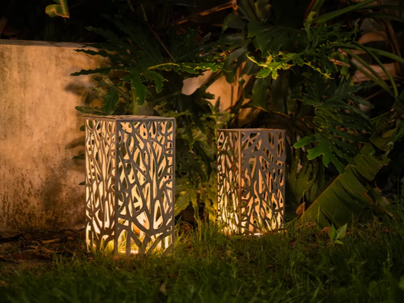 Two cylindrical laser cut aluminium garden lamps glowing among plants at night garden light idea