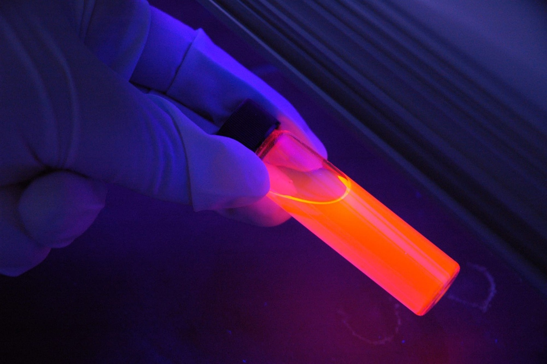 A gloved hand holds a glass vial glowing bright orange under a purple UV blacklight, showing fluorescence invisible UV light triggers